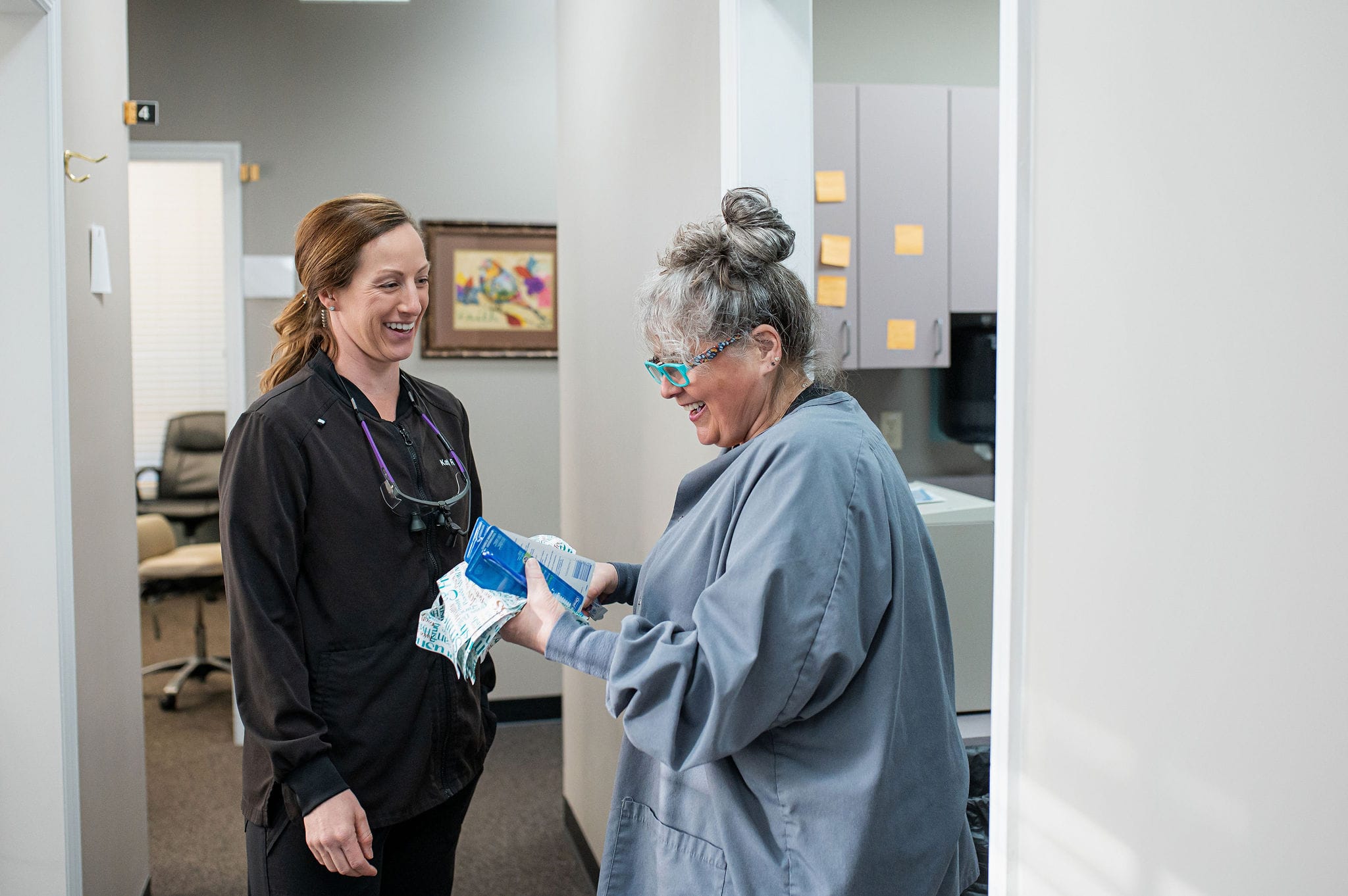 Two dental professionals are happily engaged in a conversation in a dental office hallway. One is holding dental supplies and both are smiling and laughing, creating a positive and welcoming atmosphere. The office setting includes cabinets, a framed picture on the wall, and post-it notes, suggesting an organized and friendly work environment.