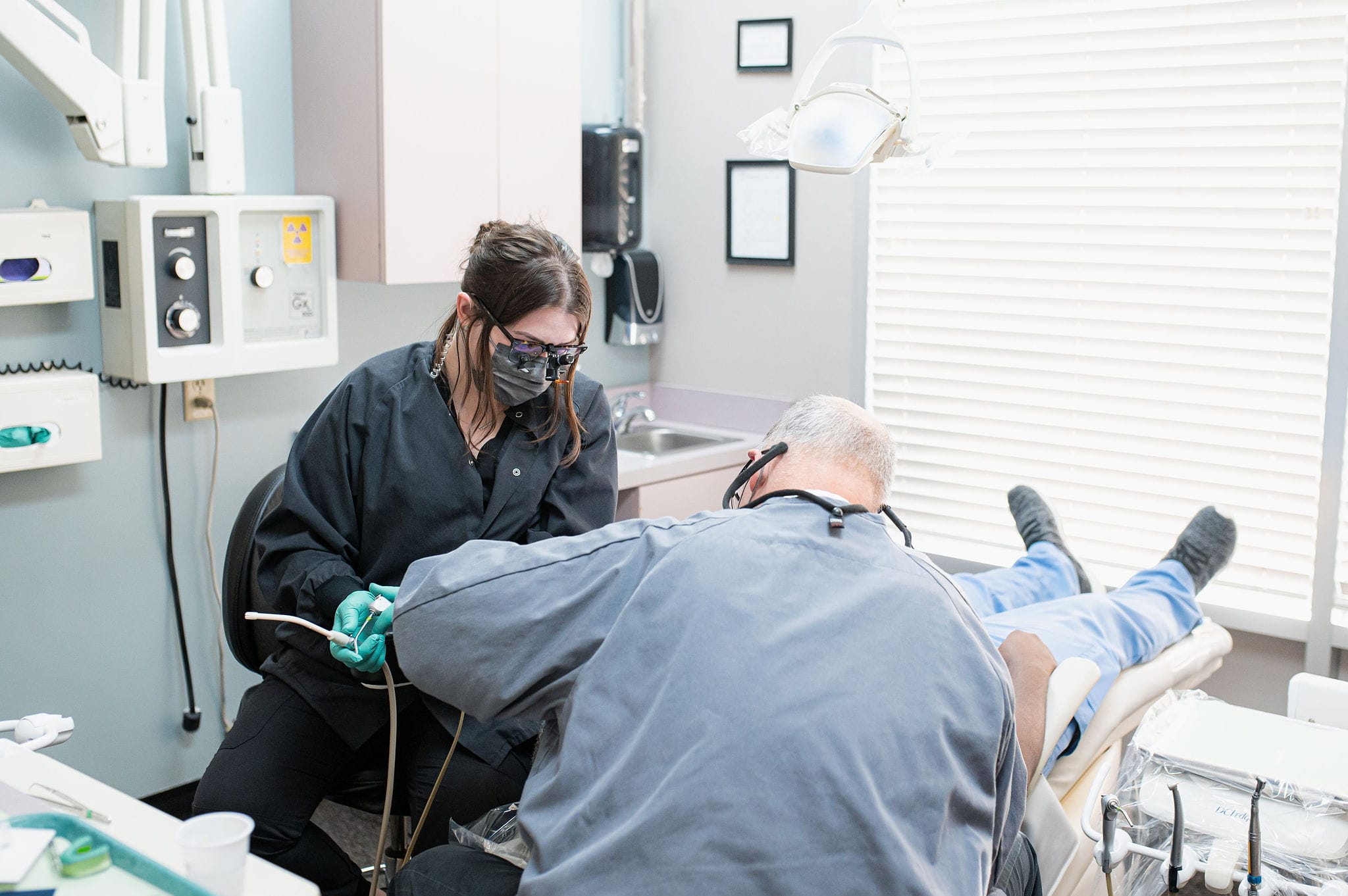 A dentist and dental assistant wearing protective gear and magnifying eyewear performing a dental procedure on a patient reclining in a dental chair, surrounded by dental equipment and supplies.