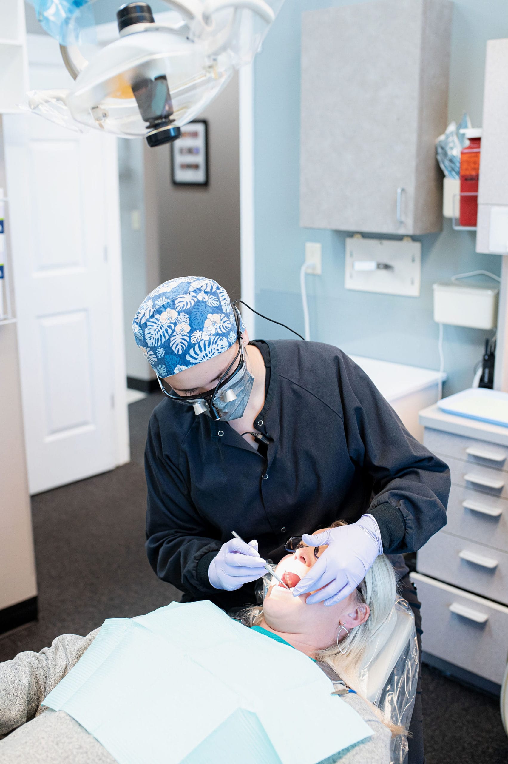 A dental professional wearing a patterned surgical cap and magnifying glasses performs a dental procedure on a patient reclining in a dental chair, with medical equipment visible in the background.