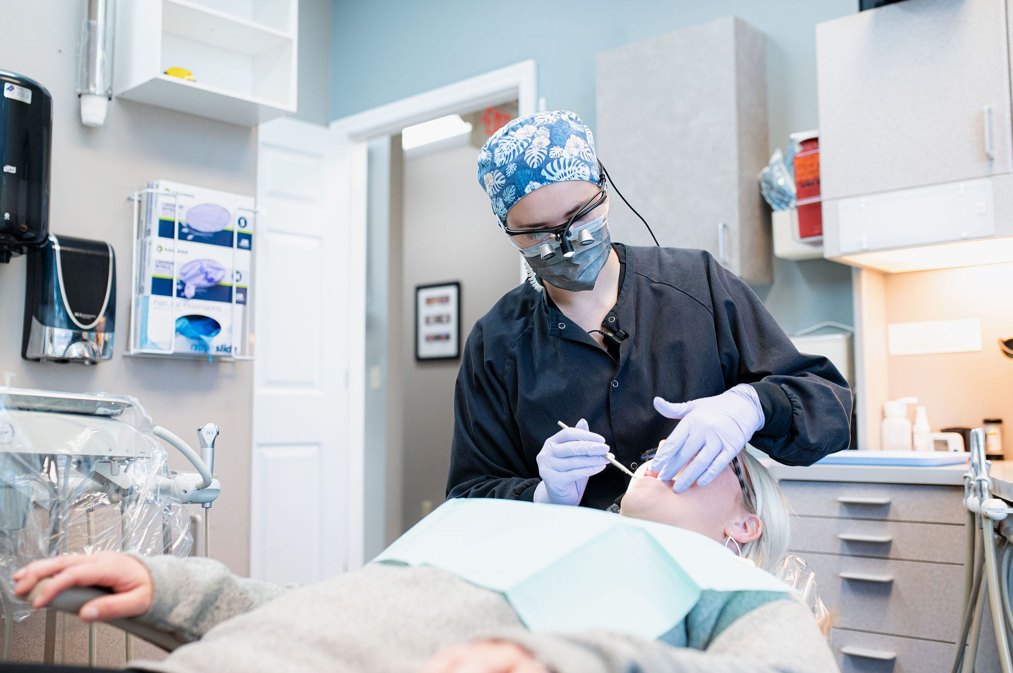 ChatGPT A dental professional in a surgical cap, mask, and magnifying eyewear performing a procedure on a patient lying in a dental chair, surrounded by dental equipment and supplies.
